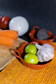Close-up of two limes, garlic, two peeled carrots, and more in clay pots with black background. Fresh vegetables in small pots above colorful handmade palm table mat. Healthy food preparation