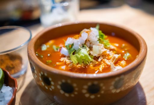 Close-up of clay bowl of delicious and traditional red pozole on wooden table. Authentic Hispanic pork stew in handmade bowl with chopped onion and lettuce on top. Traditional Mexican cuisine