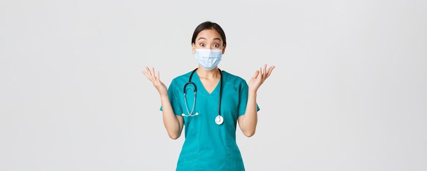 Covid-19, coronavirus disease, healthcare workers concept. Surprised and amazed asian female doctor in medical mask and scrubs, looking impressed, raising hands up, find out good news.