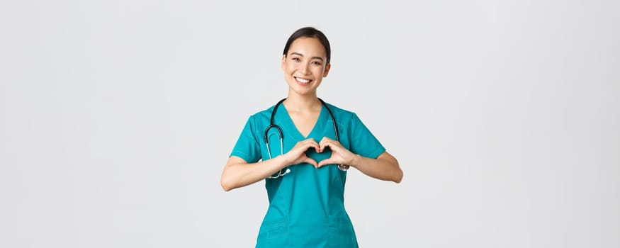 Covid-19, healthcare workers, pandemic concept. Lovely caring asian doctor, female nurse in scrubs showing heart gesture and smiling, taking care of patients with love, white background.