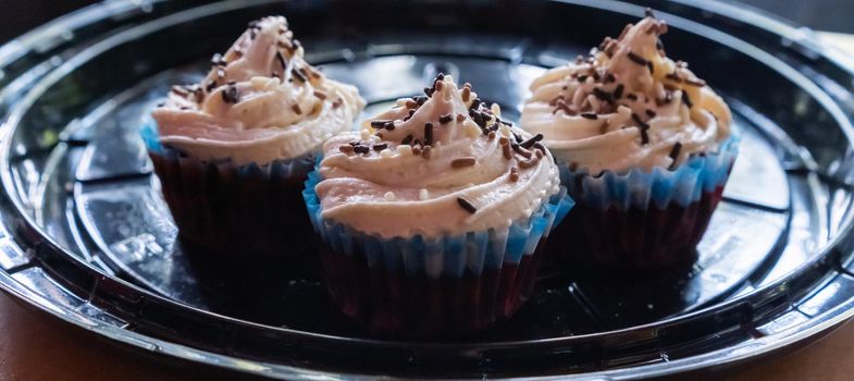 Wide view of three delicious cupcakes on round black plastic tray. Cute pastries with chocolate frosting and chocolate sprinkles above black surface. Desserts and sweet food