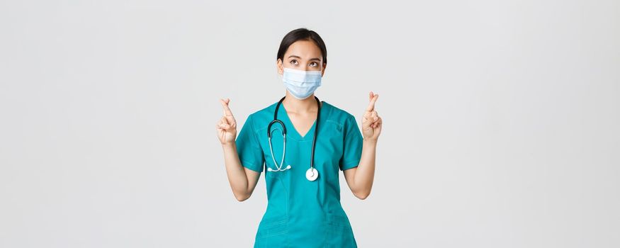 Covid-19, coronavirus disease, healthcare workers concept. Hopeful serious-looking asian doctor, female physician in medical mask and scrubs cross fingers, looking upper left corner.
