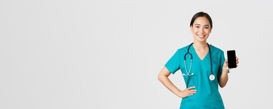 Covid-19, healthcare workers and online medicine concept. Professional female asian doctor, nurse in scrubs showing smartphone screen and smiling, showing internet appointment app, white background.