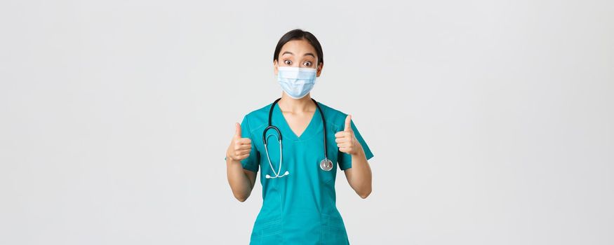 Covid-19, coronavirus disease, healthcare workers concept. Impressed happy asian female doctor, physician in medical mask and scrubs show thumbs-up, agree, compliment choice, praise nice work.