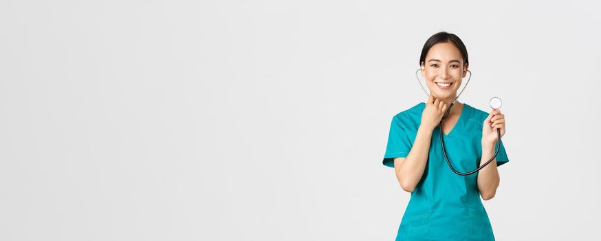 Covid-19, healthcare workers and preventing virus concept. Cheerful smiling asian female physician, doctor consult patient, examine person with stethoscope, standing white background.