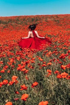 Young woman stands with her back in a long red dress and hat, posing on a large field of red poppies.