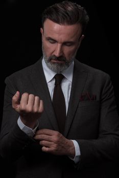 Portrait of a stylish elegant senior businessman with a beard and casual business clothes in photo studio isolated on dark background adjusting the suit