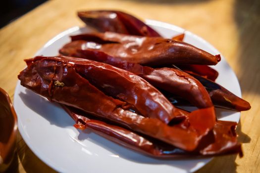 Close-up of group of dry red chili peppers on white plate above table. Several thin peppers on porcelain plate above wooden surface. Traditional hot sauce preparation