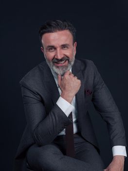 Portrait of adult businessman wearing trendy suit and sitting in modern studio on stylish chair against the black background. Horizontal mockup. High quality photo