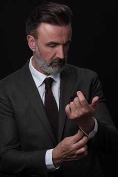 Portrait of a stylish elegant senior businessman with a beard and casual business clothes in photo studio isolated on dark background adjusting the suit