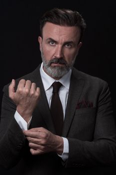 Portrait of a stylish elegant senior businessman with a beard and casual business clothes in photo studio isolated on dark background adjusting the suit