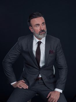 Portrait of adult businessman wearing trendy suit and sitting in modern studio on stylish chair against the black background. Horizontal mockup. High quality photo