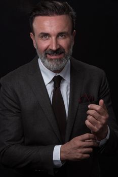 Portrait of a stylish elegant senior businessman with a beard and casual business clothes in photo studio isolated on dark background adjusting the suit