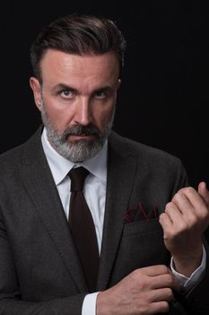 Portrait of a stylish elegant senior businessman with a beard and casual business clothes in photo studio isolated on dark background adjusting the suit