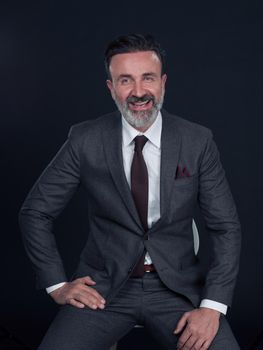 Portrait of adult businessman wearing trendy suit and sitting in modern studio on stylish chair against the black background. Horizontal mockup. High quality photo