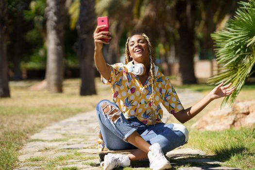 African girl with braided hair taking a selfie with a smartphone sitting on her skateboard in the street.