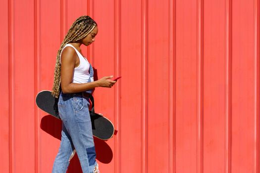 Young black girl with coloured braids walking on an urban red background while consulting her smartphone.