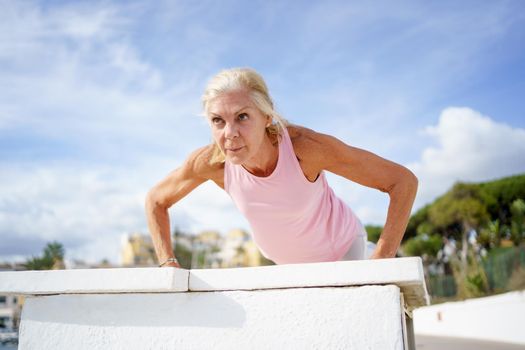 Push ups or press ups exercise by senior woman. Mature female working strength training against sky with copyspace.