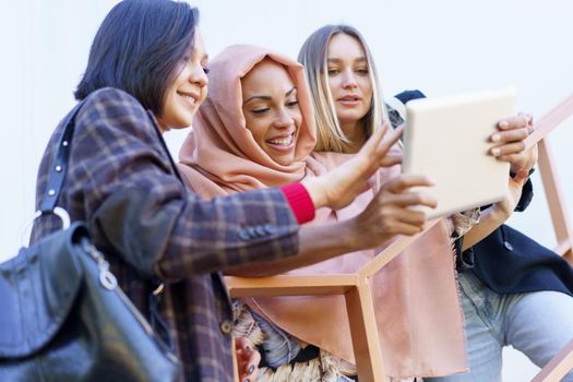 Confident young multiracial female girlfriends smiling and sharing tablet standing on metal staircase on street