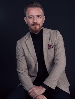 Portrait of adult businessman wearing trendy suit and sitting in modern studio on stylish chair against the black background. Horizontal mockup. High quality photo