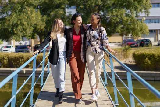 Full body of cheerful multiracial girlfriends in stylish clothes walking on footbridge over canal and looking at each other