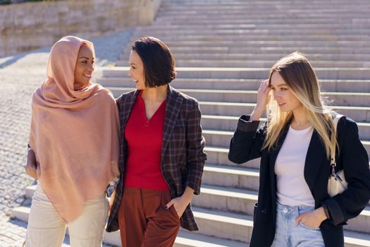 Confident young happy multiracial female students in casual outfits and hijab smiling while walking downstairs in university campus on sunny day