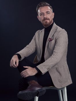 Portrait of adult businessman wearing trendy suit and sitting in modern studio on stylish chair against the black background. Horizontal mockup. High quality photo