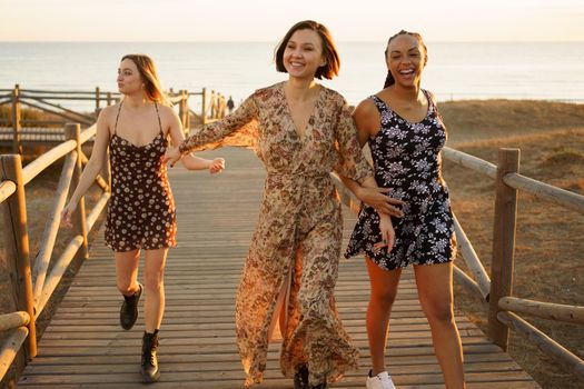 Happy multiracial women holding hands and reaching hand to girlfriend while strolling on wooden walkway on seashore at sunset