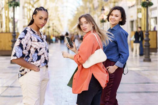 Stylish young lady with blond hair using smartphone and looking at camera while standing on street with multiethnic female friends during weekend in city
