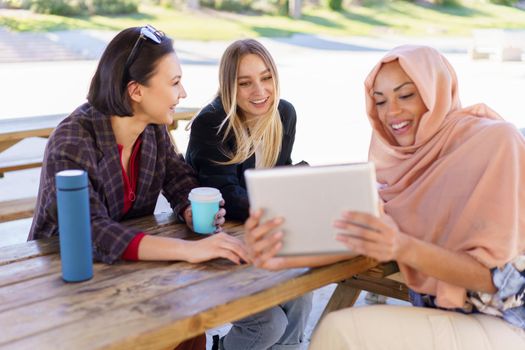Group of positive young multiethnic female friends smiling and drinking coffee, to go while watching funny video on tablet, during break in outdoor cafe in park