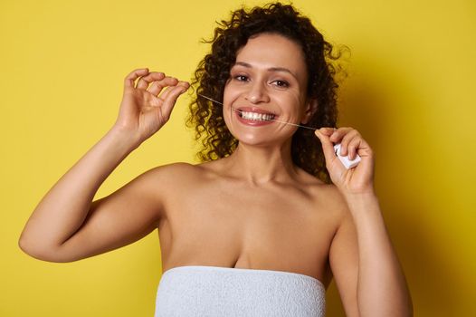 Smiling young woman taking care about her teeth by using a dental floss for cleaning dental cavity, isolated over yellow background with space for text