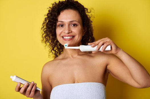 A mixed race woman holding a toothbrush and toothpaste ready to brush her teeth looking at the camera isolated over yellow background with space for text