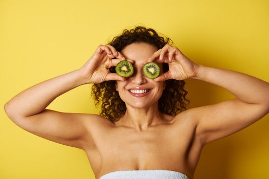 Smiling mixed race muscular woman covering her eyes with kiwi halves, smiling while posing against yellow background with space for text
