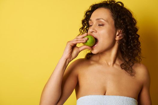 A curly-haired swarthy woman bites a green apple while standing on a yellow background. Healthy lifestyle concepts