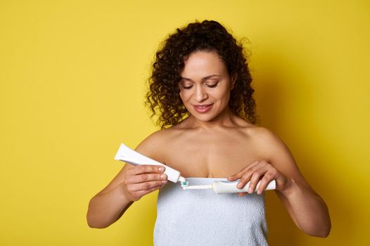 Young mixed race woman wrapped in white towel squeezing a toothpaste on an electric toothbrush