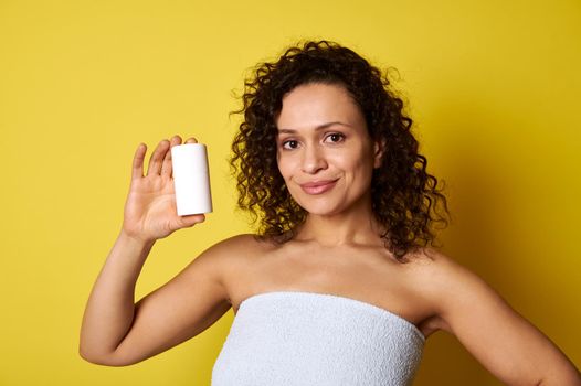 Beautiful smiling middle aged woman holding cosmetic product bottle isolated on yellow background with copy space. Attractive curly mixed race young woman posing with bottle.