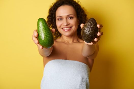 Soft focus on ripe ready-to-eat avocado fruit in hands of blurry half-naked curly woman wrapped in towel against yellow background