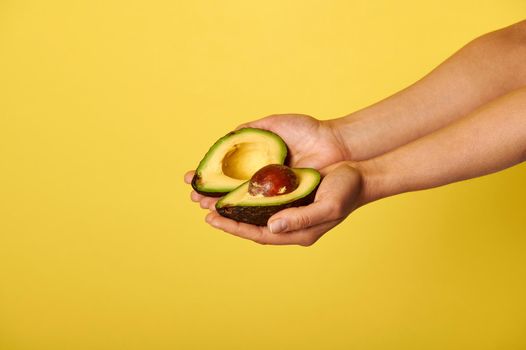 Closeup of hands holding two halves of ripe ready-to-eat avocado. Closeup on yellow background with copy space