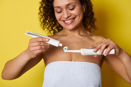 Closeup of a smiling African woman squeezing toothpaste on electric toothbrush, isolated over yellow background with copy space