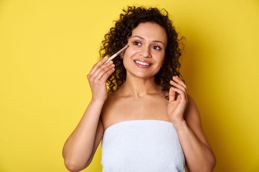 Beautiful mulatto woman applying decorative cosmetics with makeup brush and smiling while posing for camera on yellow background with space for text