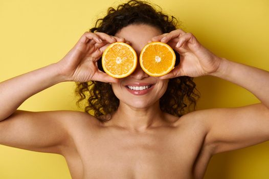 Smiling mixed race muscular woman healthy smile and glowing hydrated facial skin, covering her eyes with sweet orange halves, smiling while posing against yellow background with space for text