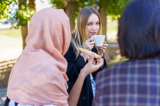Young lady with long blond hair drinking coffee and talking with anonymous diverse female friends in outdoor cafe on sunny day