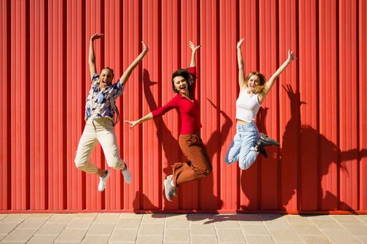 Full length of cheerful young multiracial female best friends in casual clothes shouting and jumping against metal red wall on city street