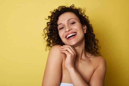 Beauty portrait of young half naked woman with curly hair smiling toothy smile looking at camera, posing over yellow background with copy space