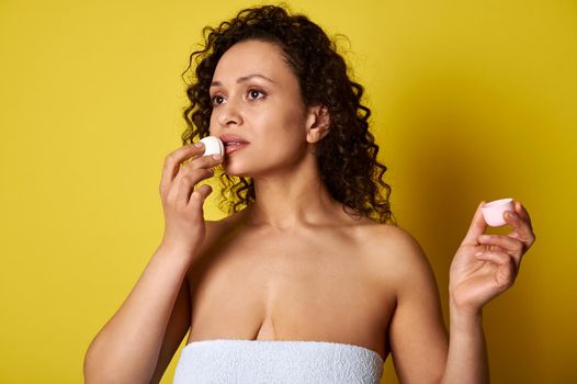 A young woman wrapped in a bath towel using a hygienic lipstick for hydrating and taking care about her lips. Copy space, yellow background