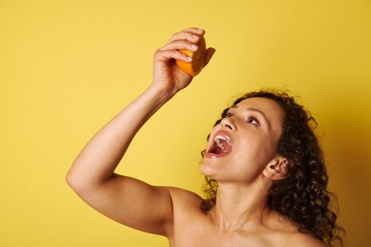 Beautiful half naked woman squeezing orange juice into her mouth. Closeup isolated on yellow background with copy space
