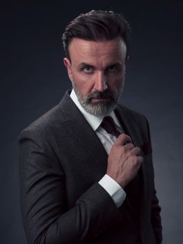 Portrait of a stylish elegant senior businessman with a beard and casual business clothes in photo studio isolated on dark background adjusting the suit