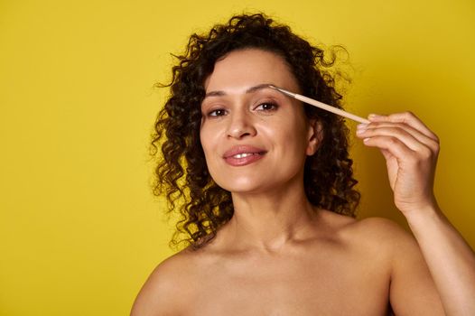 Smiling dark skin woman using makeup brush to correct and dye eyebrows. Make-up concepts on yellow background with copy space