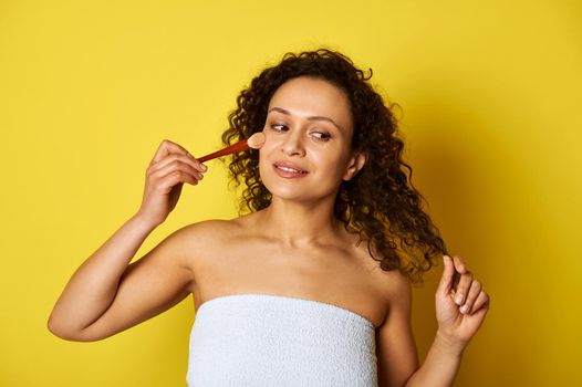 Charming half naked mulatto woman, wrapped in white bath towel, holding a makeup brush and applying blush on her face. Isolated beauty concepts on yellow background with copy space