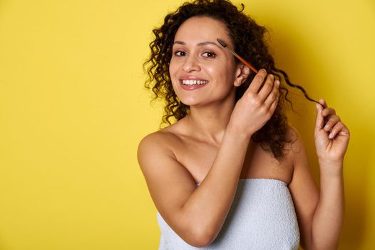 Smiling mixed race young woman with perfect skin and makeup posing with makeup brush over yellow background with space for text